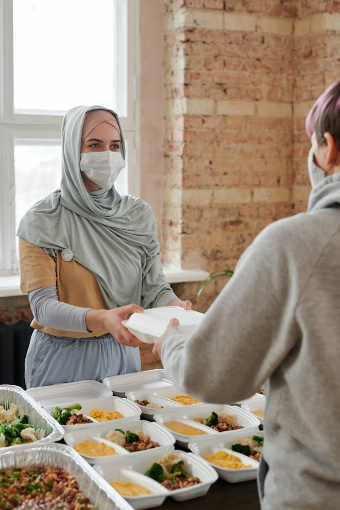 A woman wearing a hijab and face mask hands out packed food indoors, promoting charity and safety.